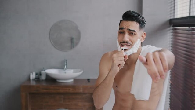 Cheerful funny goofy handsome arab man guy with white soap suds on beard naked male with towel on shoulder singing song into razor having fun shaving in bathroom at home dancing music humorous hygiene