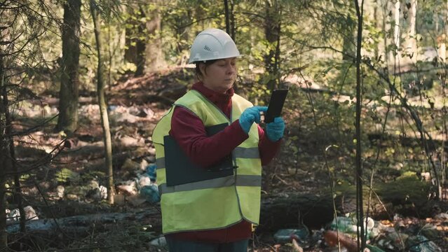 Woman environmentalist in workwear and helmet record offense - forest pollution and issue a fine. Concept of pollution of natural parks, woods and nature reserves with non decomposable waste