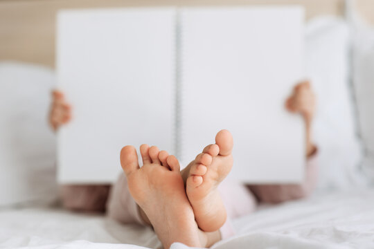 Children bare feet and hands close up. Kid reading  book in white cozy bed before sleepping night. Copy space, mock up,plase for text. Rest and holiday in hotel