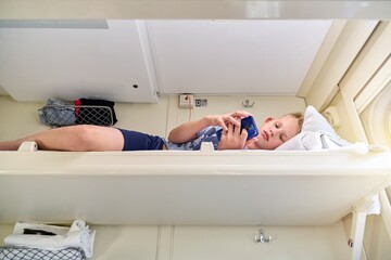 Teen boy with short blond hair reads electronic book with smartphone lying on white bedding on...