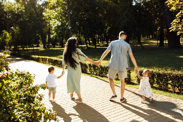 Stylish parents with small children walking at park, enjoying joyful moments together. Loving father with caring mother holding hands with little kids, spending weekends outdoors, happy family concept