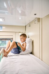 Blond little boy watches cartoons via tablet PC and leaning on soft pillow on top shelf in train...