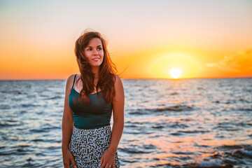 A young charming woman in a skirt stands on the seashore in front of a bright orange sunset. Romantic seascape