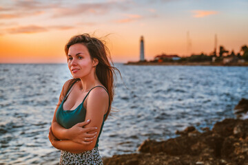 A romantic young woman in a skirt admires the sunset on the sea with a background in the form of a lighthouse in the Crimea