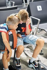 Concentrated blond brothers play games and share interests using smartphones and sitting in waiting hall with armchairs in airport