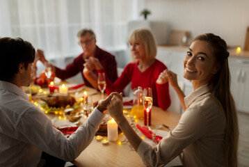 Young woman celebrating Christmas with her husband and senior parents, having festive Xmas dinner, praying to God
