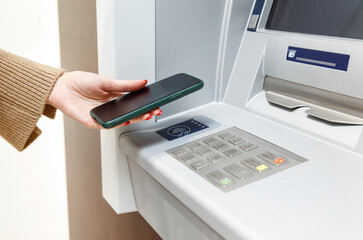 Female hand with mobile phone withdrawing money from atm using NFC contactless wi-fi pay pass system. - 484911942