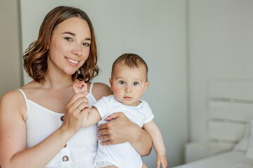 A beautiful young mother holds her little brown-eyed 8 month old daughter in her arms. Portrait of a family in a beautiful cozy house. Motherhood. Mothers Day.
