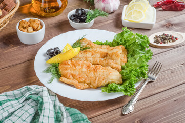Fried fish fillet pieces with lemon and herbs on a white plate on a dark wooden background