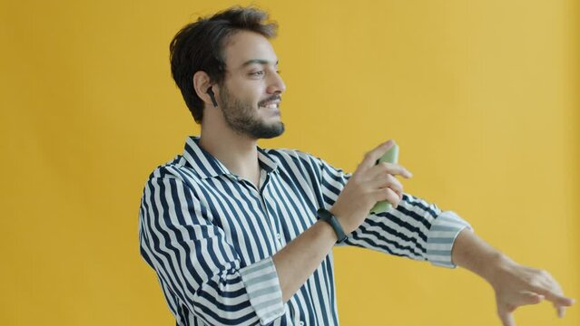 Portrait of Indian guy using earpods and smartphone dancing having fun on yellow color background. Modern devices and entertainment concept.
