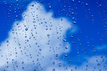 blue rain water drops on a window glass close up with white colorful clouds in background, drop and cloud macro texture ; bad weather backdrop