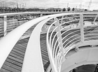 A white metal handrail curves in an arc against the backdrop of a marina with yachts and palm trees. Black and white photo