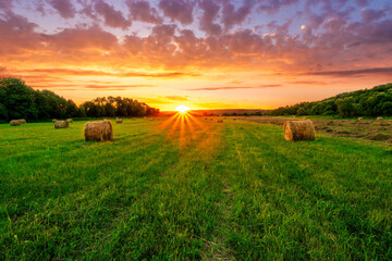 Fototapeta premium Scenic view at picturesque burning sunset in a green shiny field with hay stacks, bright cloudy sky , trees and golden sun rays, summer valley landscape