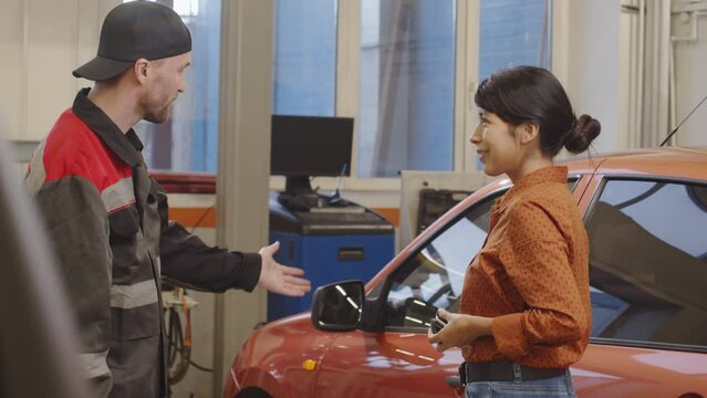 Professional mechanic in uniform giving car key to female client and talking with her in auto service garage