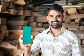 happy smiling Indian artisan with green screen mobile phone looking at camera at carpentry shop - conept of online booking of woodworking service, advertising and blue collar jobs