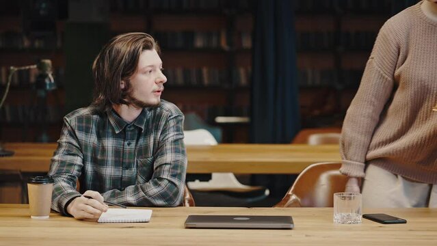 Young pensive man studying with notebook at library, woman groupmate coming with books and giving one to him