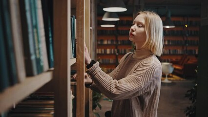 Young woman student coming to bookshelves in library and choosing book, reading information, tracking shot