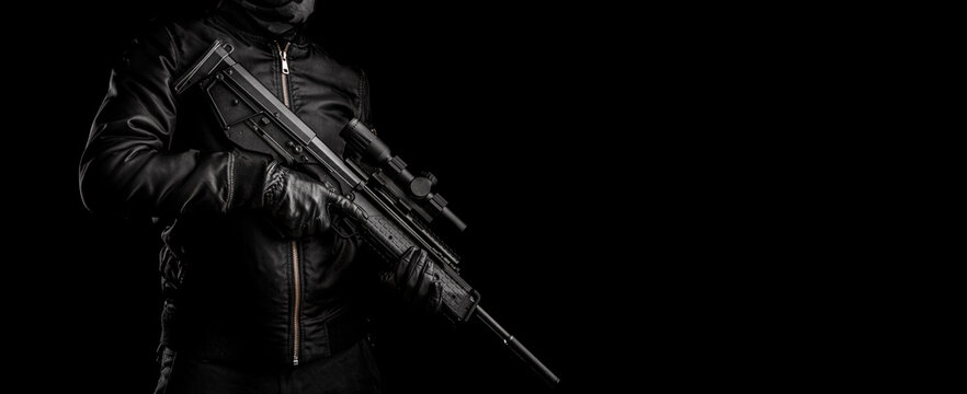 A man in dark clothes holds an automatic carbine in his hands. The face is covered with a mask, hands are in gloves. A figure with a weapon on a dark back