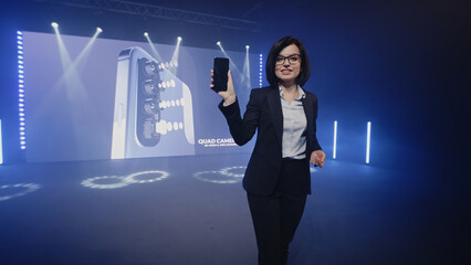 Female host in a suit on stage showing a new fashionable device, during the presentation of the release near the LED screen with a 3D mock-up in a large illuminated room