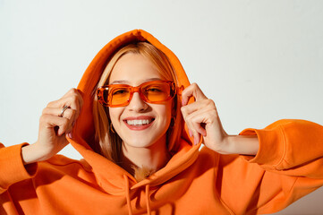 Happy smiling fashionable woman wearing trendy orange color sunglasses, hoodie posing on white...