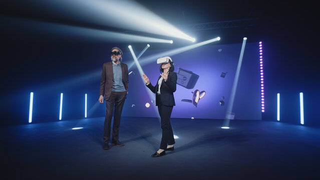 Two speakers in virtual reality glasses standing on the stage and talking about modern technologies and the development of the Metaverse, during presentation in illuminated room with an LED screen