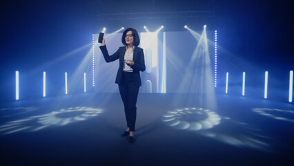 Female host in a suit on stage showing a new fashionable device, during the presentation of the release near the LED screen with a 3D mock-up in a large illuminated room