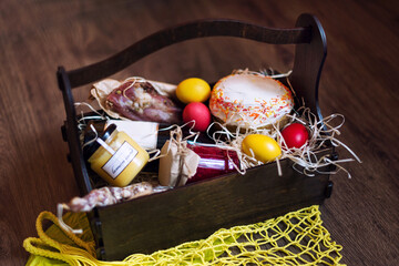 Colorful easter eggs in a basket with cake, red wine, hamon or jerky and dry smoked sausage on wooden background. - Powered by Adobe