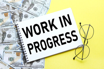 Work in progress text on an open copybook with a spiral on the background of money scattered on the table