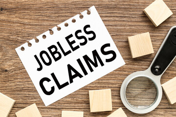 Jobless claims text on white paper from a notepad on a wooden background. wooden blocks magnifier