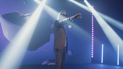 Developer in a casual suit on stage presenting and talking about new virtual reality headset for the Metaverse, conference in a dark lit room opposite the LED screen with 3D objects