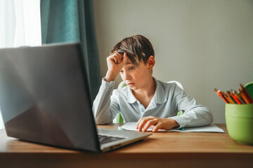 boy is sitting at his desk, in front of him is a notebook and a laptop, distance learning, a student is doing homework, the child is upset, does not understand a difficult task