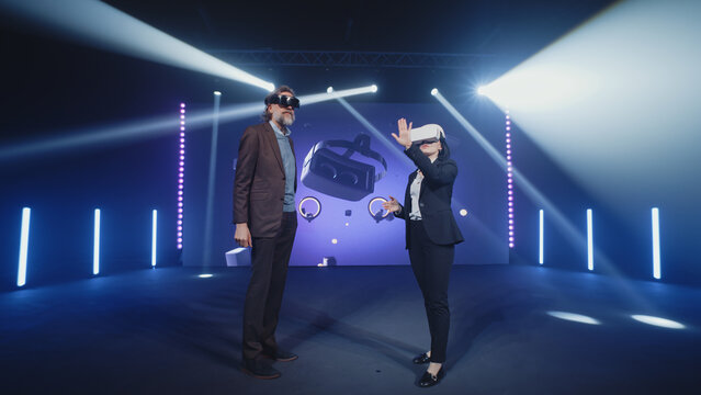 Two speakers in virtual reality glasses standing on the stage and talking about modern technologies and the development of the Metaverse, during presentation in illuminated room with an LED screen