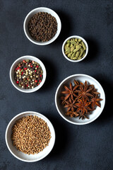 Spices assorted multicolor peppercorns, cardamom, star anise, cumin, coriander, caraway in bowls on black background.