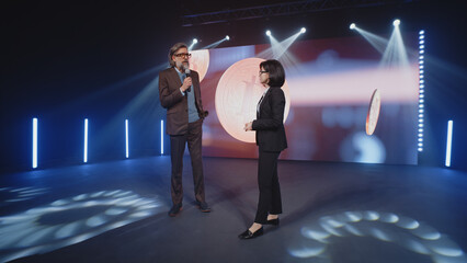 A female presenter in a suit interviewing a cryptocurrency coach on stage in front of an LED screen...