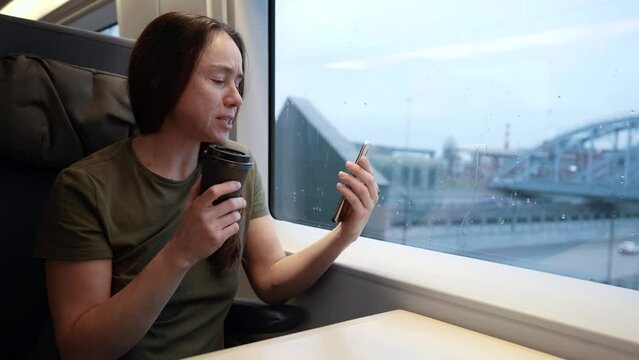 An adult woman travels by train and communicates via the Internet via video link. A lady on the train is sitting at a table with a disposable cup of coffee.
