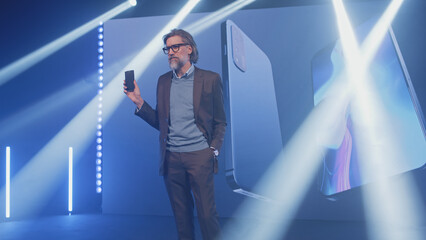 Male adult creator in a casual suit on stage presenting a modern smartphone with new technologies...