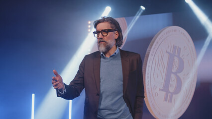 Focused speaker in a casual suit and glasses on stage, gesturing and speaking about bitcoin and...