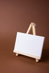 White canvas on a small wooden easel stand minimal concept on the brown backdrop. Empty blank mock up frame for design with copy space.
