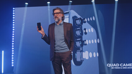 Male adult creator in a casual suit on stage presenting a modern smartphone with new technologies to the audience in front of an LED screen, with a 3D mock-up in a lit room during a release event