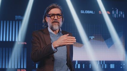 A mature businessman in a suit on stage telling the audience about the financial success of company and investments in front of an LED screen with graphs, during the presentation of business strategy