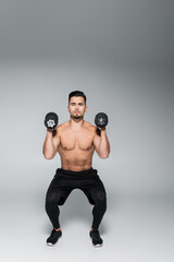 full length of strong sportsman doing squat with dumbbells on grey.