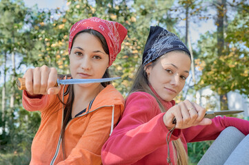 Two young college backpacker women take a break, back to back, look to the camera showing their knives.  - obrazy, fototapety, plakaty