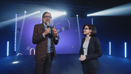 A female presenter interviewing a male developer of a new augmented reality device during the presentation of meta universe in a modern studio with spotlights, near the LED screen in 3D objects - obrazy, fototapety, plakaty