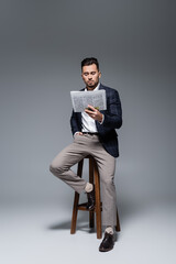 full length of bearded businessman in checkered blazer sitting on chair and reading newspaper on grey.