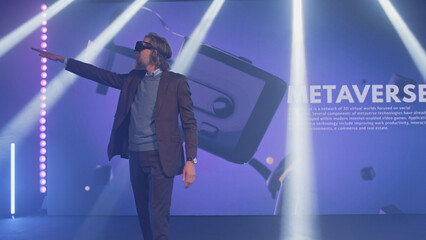 Happy male speaker in suit gesturing and demonstrating augmented reality glasses, to the audience at a Metaverse event on stage in front of an LED screen 3D objects in illuminated room