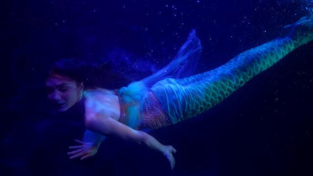 romantic and dramatic mermaid is swimming in dark blue depth of lake or sea, slow motion underwater