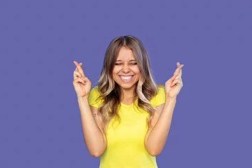 Papier Peint photo Pantone 2022 very peri A young pretty smiling blonde woman in a yellow t-shirt with her eyes closed crosses her fingers for good luck waiting for the results of the lottery or exams isolated on a very peri color background