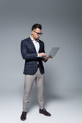 Fototapeta na wymiar full length of businessman in suit and glasses using laptop on grey.