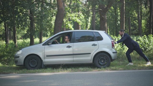 Young Caucasian woman sitting on driver's seat as man pushing vehicle outdoors on sunny day. Wide shot couple with broken automobile on suburban road trying start the car