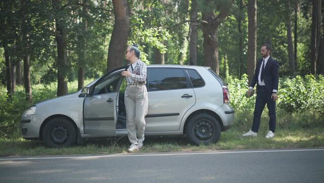 Wide shot woman walking out driver's seat helping man pushing vehicle on suburban roadside on sunny day. Young Caucasian couple with broken automobile outdoors. Transport and lifestyle